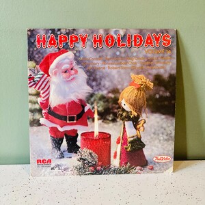 May include: A vinyl record cover titled Happy Holidays Volume 14 featuring a Santa Claus figurine and a doll standing near a lit red candle. The text includes names of performers, the RCA Special Products logo, and the True Value hardware store logo.