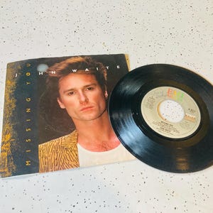 May include: A 7-inch vinyl record and its paper sleeve featuring John Waite. The sleeve displays the text "MISSING YOU" and "JOHN WAITE" alongside a portrait of a person in a yellow patterned jacket. The black record has a central label with text.