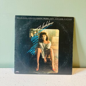 May include: A black vinyl record album cover with the title "Flashdance" in blue and white lettering. The cover features a woman in a white shirt and red shoes sitting on a stone ledge.