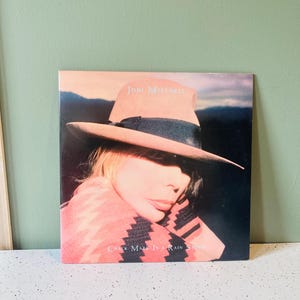 May include: Album cover for Joni Mitchell's "Chalk Mark in a Rain Storm." The cover features a portrait of Mitchell wearing a hat, with the album title and artist's name displayed. The color palette includes pink, black, and shades of brown.