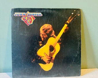 May include: Vintage vinyl record album cover featuring John Denver playing a yellow acoustic guitar. The album cover is black with the artist's image and the text "JOHN DENVER" in a colorful logo.