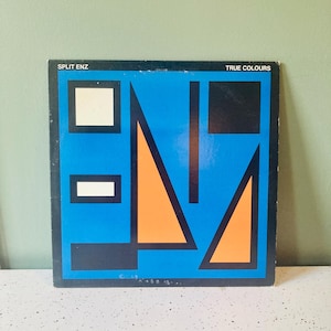 May include: A square vinyl record cover for Split Enz titled True Colours. The cover features a bright blue background with black and orange geometric shapes, including triangles and rectangles. The text Split Enz and True Colours is printed in white.