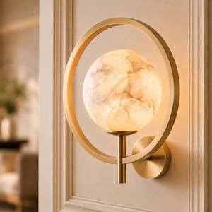 May include: A wall-mounted light fixture featuring a circular brass frame surrounding a spherical, glowing white orb with a marbled texture. The lamp is attached to the wall by a circular brass base, creating a balanced and geometric appearance.