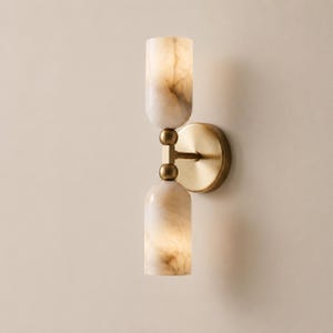 May include: A wall-mounted light fixture featuring a circular brass backplate and a central brass arm. Two cylindrical, translucent stone shades with marbled beige and white patterns are positioned vertically, one above the other, on the arm.