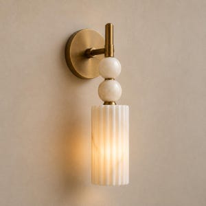 May include: A wall sconce featuring a circular brushed brass backplate and a vertical arm. The fixture includes two stacked white marble spheres and a cylindrical, vertically fluted white marble shade that emits a warm, soft internal glow.