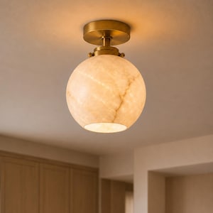 May include: A flush mount ceiling light featuring a spherical, translucent stone shade with natural veining patterns. The fixture includes a circular brass-toned metal canopy and a matching hardware mount that secures the globe to the ceiling.