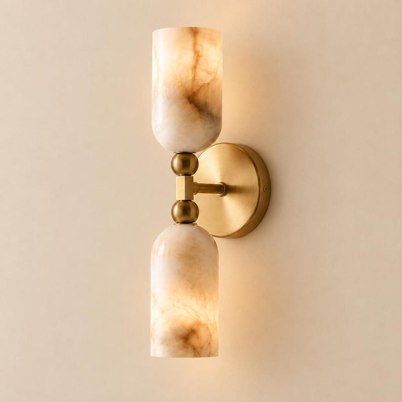 May include: A wall sconce featuring a circular brushed brass backplate and a central vertical arm. Two cylindrical, translucent, marbled stone shades are positioned at the top and bottom, each secured by a small brass sphere connector.
