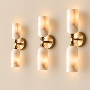 May include: Three wall sconces featuring cylindrical, cream-colored marble shades with brown veining. Each fixture has a central brass-toned metal arm with two spherical accents connecting the top and bottom shades to a circular wall-mounted base.