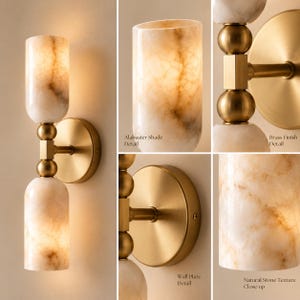 May include: A wall sconce featuring two cylindrical alabaster shades with natural veining and a brushed brass finish. The fixture includes a circular wall plate, a central brass stem with spherical accents, and illuminated stone shades.