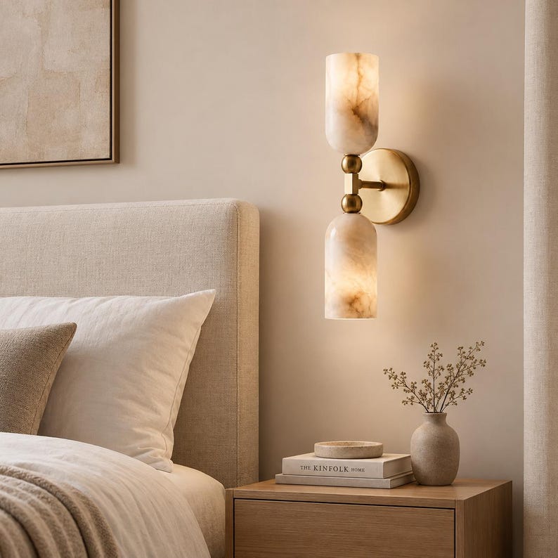 May include: A wall-mounted light fixture featuring two cylindrical, translucent stone shades with marbled beige and white patterns. The shades are connected by a central brass-toned metal rod and circular wall plate, emitting a warm, soft glow.