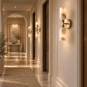May include: A wall-mounted light fixture featuring two cylindrical, marbled, off-white glass shades connected by a central brass-toned metal arm. The fixture is attached to a circular brass-toned base plate and emits a warm, soft glow.