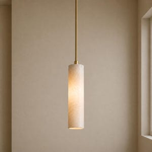May include: A cylindrical pendant light fitting featuring a translucent, cream-coloured stone shade with subtle veining. The shade is suspended from a thin, vertical brass-toned metal rod that extends upwards, creating a sleek and elongated profile.