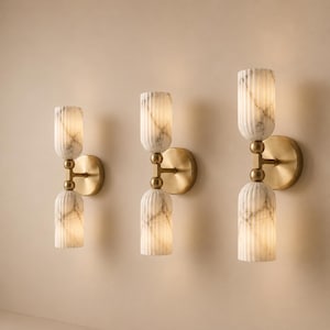 May include: Three wall sconces featuring a brass circular base and a central brass rod connecting two vertically stacked, fluted, white marble-patterned glass shades. Each fixture is mounted to the wall, showcasing a symmetrical design.
