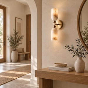 May include: A wall-mounted light fixture featuring two cylindrical, marbled stone shades in a warm cream tone, connected by a central brass-finished metal arm. The fixture emits a soft, diffused glow from both the top and bottom glass cylinders.