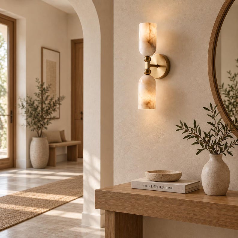 May include: A wall-mounted light fixture featuring two cylindrical, marbled stone shades in a warm cream tone, connected by a central brass-finished metal arm. The fixture emits a soft, diffused glow from both the top and bottom glass cylinders.
