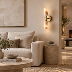 May include: A wall-mounted light fixture featuring two cylindrical, translucent, cream-colored stone shades with brown veining, connected by a brass-toned metal arm and a circular wall plate, emitting a warm, soft glow in a neutral-toned room.