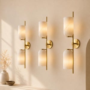 May include: Three wall-mounted light fixtures, each featuring a vertical brass-toned metal rod supporting two cylindrical, vertically ribbed white glass shades. The fixtures are mounted to the wall with circular brass-toned metal plates.