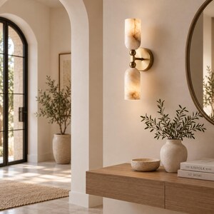 May include: A wall-mounted light fixture featuring two cylindrical, translucent stone shades with marbled beige patterns, connected by a central brass-toned metal arm and a circular wall plate, emitting a warm, soft glow against the wall.