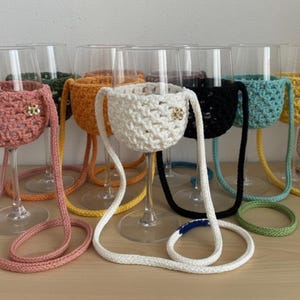 Könnte beinhalten: Eine Sammlung von Weingläsern aus klarem Glas, die jeweils mit einem abnehmbaren, gehäkelten Garnhalter in verschiedenen Farben wie Rosa, Orange, Weiß, Schwarz, Türkis und Gelb ausgestattet sind, inklusive langer passender Trageriemen und kleiner Metallakzente.