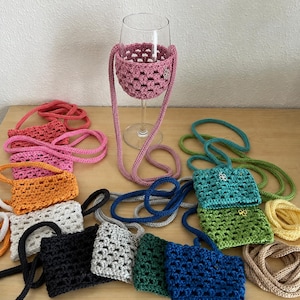 Könnte beinhalten: Eine Sammlung von gehäkelten Weinglashaltern mit langen Trägern in verschiedenen Farben wie Pink, Orange, Weiß, Schwarz, Grau, Grün und Blau. Ein pinker Halter ist um ein klares Glas gelegt, während andere flach auf einer Oberfläche liegen.