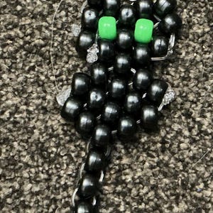 May include: A keyring featuring a cat figure constructed from black plastic pony beads with two bright green beads for eyes. The cat figure is attached to a silver-coloured metal star-shaped clasp by a small metal jump ring and chain link.