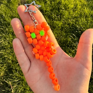 May include: A keyring crafted from bright orange plastic pony beads shaped into a small animal figure with two neon green bead eyes. The figure features a long tail and is attached to a silver-coloured metal star-shaped split ring clasp.