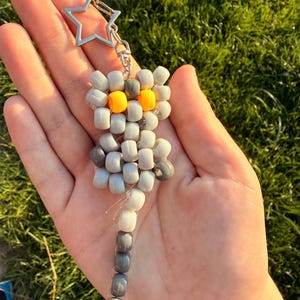May include: A keyring featuring a small animal figure crafted from grey and white plastic pony beads with two bright yellow beads for eyes. The figure is attached to a silver-toned metal star-shaped clasp by a short silver metal chain link.