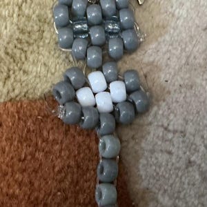 May include: A keyring crafted from grey and white plastic pony beads arranged in the shape of a cat. The design features a grey head with two ears, a grey and white body, and a long grey tail ending in a single white bead. A silver star-shaped clasp is attached to the top of the head.