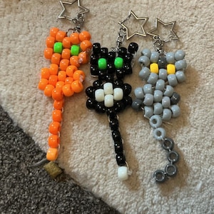May include: Three cat-shaped keychains crafted from plastic pony beads. One is orange with neon green eyes, one is black with neon green eyes and a white chest, and one is grey with yellow eyes. Each features a silver-toned star-shaped keyring.