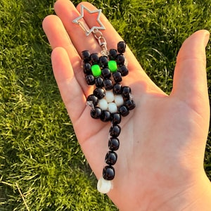 May include: A cat-shaped keyring crafted from black plastic pony beads, featuring two bright neon green beads for eyes and a cluster of white beads on the chest. The figure includes a long tail and is attached to a silver-toned metal star-shaped clasp.