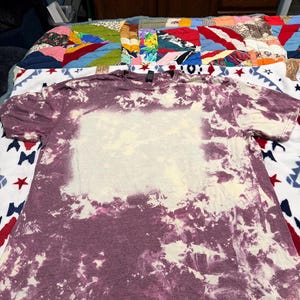 May include: A short-sleeved crew neck t-shirt featuring a purple and white bleached tie-dye pattern. The garment has a large, rectangular, light-colored bleached area in the center of the chest, surrounded by a mottled purple and white design.