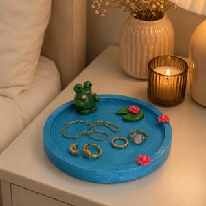May include: A round blue tray featuring a small green frog figurine, a green lily pad with a pink flower, and two additional pink flower accents. The tray holds a gold chain necklace and four gold rings, including one with a white stone centre.