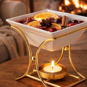 May include: A white square ceramic bowl held by a gold-toned metal stand, containing a warm liquid with orange slices, red cranberries, a star anise, and cinnamon sticks. A small lit tea light candle sits in a gold holder beneath the bowl.