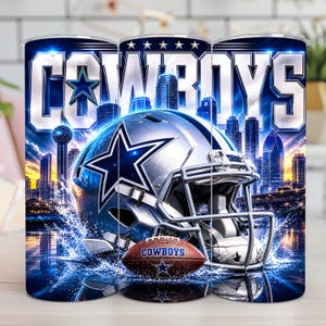 May include: A cylindrical tumbler featuring a Dallas Cowboys football helmet, a brown football with the text COWBOYS, and a city skyline background. The design includes blue and silver tones with a water splash effect at the base of the helmet.