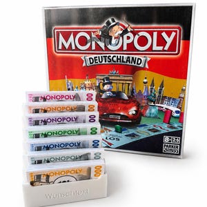 Könnte beinhalten: Ein Monopoly Deutschland Brettspielkarton mit der Figur Rich Uncle Pennybags und einem roten Auto, begleitet von einer weißen Kunststoff-Sortierbox, die sieben Stapel farbiger Monopoly-Spielgeldscheine mit dem Text Wunschtext enthält.