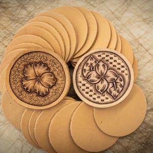 May include: A stack of round leather coasters with two coasters featuring floral designs. The coasters are a light brown colour.