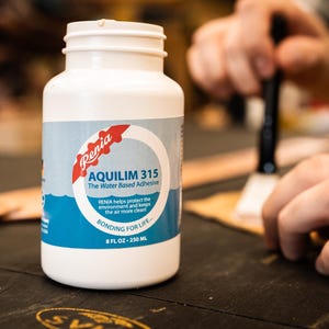May include: A white plastic bottle of Renia Aquilim 315 water-based adhesive. The label is blue and red with the text "Renia Aquilim 315 The Water Based Adhesive" and "BONDING FOR LIFE - 8 FL OZ - 250 ML".
