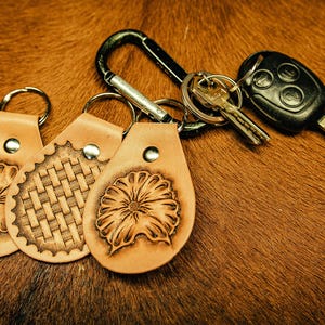 May include: Three leather keychains with different designs: a flower, a woven pattern, and another flower. The keychains are attached to a silver carabiner and a car key.