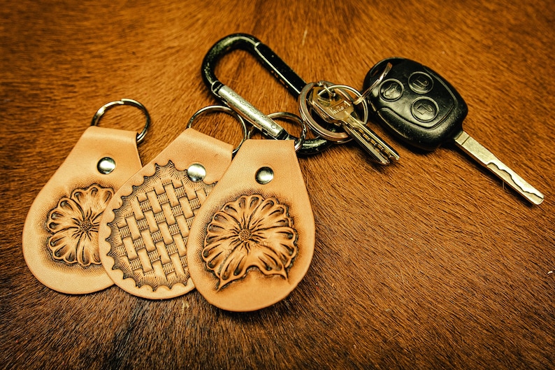 May include: Three leather keychains with different designs: a flower, a woven pattern, and another flower. The keychains are attached to a silver carabiner and a car key.
