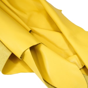 Puede incluir: Un trozo de cuero amarillo doblado y drapeado sobre un fondo blanco. El cuero tiene una superficie lisa y ligeramente texturizada.