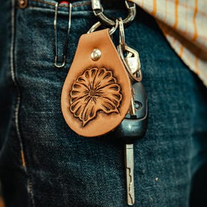May include: A brown leather keychain with a tooled floral design. The keychain is attached to a pair of keys and a silver key ring.