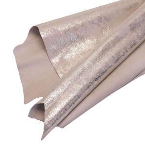 May include: Close-up of a piece of light beige leather with a metallic silver overlay. The leather is folded, showing the texture and sheen of the material. The edges of the leather are visible, highlighting the layers.