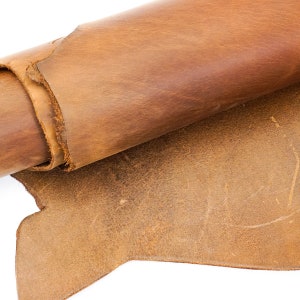 May include: Close-up of a roll of brown leather with a distressed finish. The leather has a natural, textured appearance with variations in color and tone. The edges of the leather are visible, showing the thickness and texture.