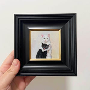 May include: A small square painting in a thick black frame with a thin gold inner border. The artwork depicts a white cat standing behind and hugging a smaller black cat. The black cat has its mouth open, showing its tongue, while the white cat looks forward.