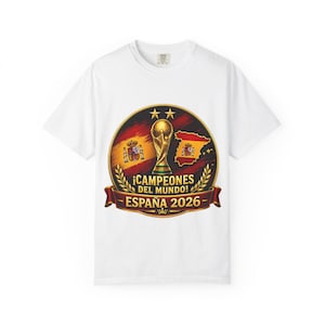 Può includere: Una t-shirt bianca con una grafica circolare raffigurante un trofeo della coppa del mondo dorato, la bandiera spagnola e una mappa della Spagna. Il testo recita ¡CAMPEONES DEL MUNDO! ESPAÑA 2026, con due stelle dorate e rami di alloro.