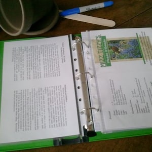May include: A green binder with a clear plastic sleeve containing a white sheet of paper with text. The text on the sheet of paper is about gardening. The binder has a silver ring mechanism and is open to reveal the contents of the sleeve. The sleeve also contains a white card with the text "Mizz Tizzys Weeds & Seeds" and a picture of purple flowers.