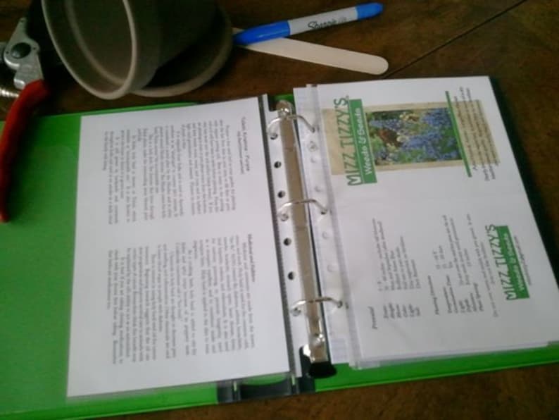 May include: A green binder with a clear plastic sleeve containing a white sheet of paper with text. The text on the sheet of paper is about gardening. The binder has a silver ring mechanism and is open to reveal the contents of the sleeve. The sleeve also contains a white card with the text "Mizz Tizzys Weeds & Seeds" and a picture of purple flowers.
