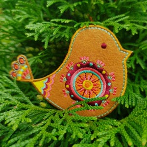 Op de afbeelding: Een vilten vogelversiering met een kleurrijk geborduurd ontwerp. De vogel is oranje met een gele, blauwe en roze staart. De borduurwerk heeft een cirkelvormig patroon met roze, oranje, gele, groene en paarse kleuren.