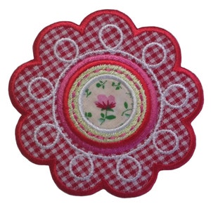 May include: A red and white gingham fabric flower with a pink and green floral centre. The flower has a scalloped edge and is surrounded by a white circle.