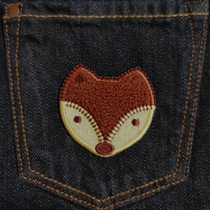 Op de afbeelding: Een geborduurde vos-patch in bruin en crème op een blauwe denim achtergrond.